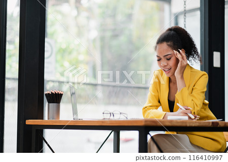 A young woman in a yellow blazer works on her laptop in a bright, modern office with large windows and natural light. 116410997