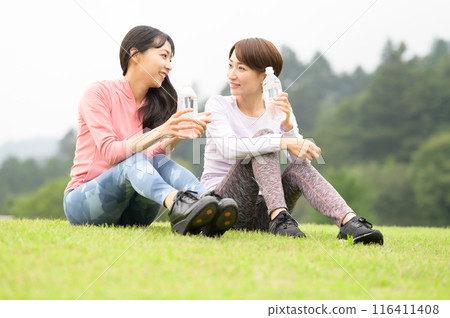 Two women hydrating on the grass after exercise Two women hydrating on the grass after exercise 116411408
