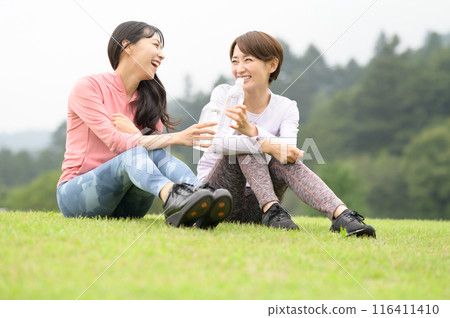 Two women hydrating on the grass after exercise 116411410
