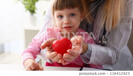 Little baby girl visiting doctor holding in hands red toy heart Little baby girl visiting doctor holding in hands red toy heart 116412923