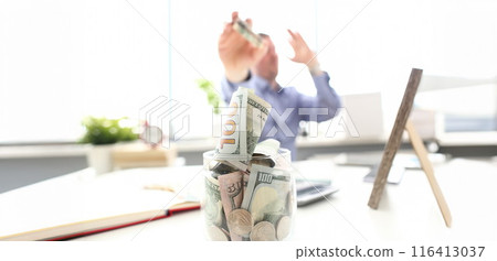 Rich man throwing US banknotes in big jar full of money at workplace 116413037