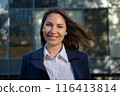 Portrait of an attractive young adult woman in a blue suit standing against the background of an office building, smiling and looking at the camera close-up 116413814
