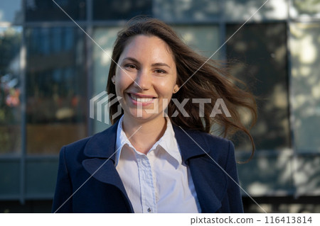 Portrait of an attractive young adult woman in a blue suit standing against the background of an office building, smiling and looking at the camera close-up 116413814