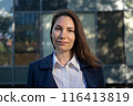 Portrait of an attractive strong confident independent woman in a blue suit standing against the background of a business building and looking at the camera 116413819