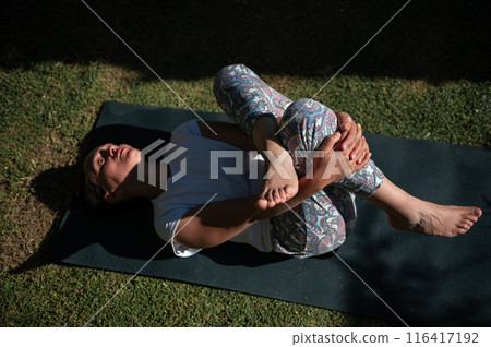 Woman practicing yoga outdoors on a sunny day, performing a relaxing pose on a yoga mat 116417192