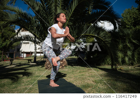 Woman practicing tree pose yoga outdoors in a serene park setting, surrounded by lush greenery Woman practicing tree pose yoga outdoors in a serene park setting, surrounded by lush greenery 116417249