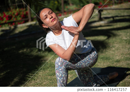 Woman practicing yoga outdoors in a park on a sunny day, performing a twisting pose 116417257