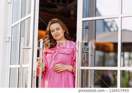Beautiful pregnant woman in pink dress standing in the doorway of the house. Beautiful pregnant woman in pink dress standing in the doorway of the house. 116417493