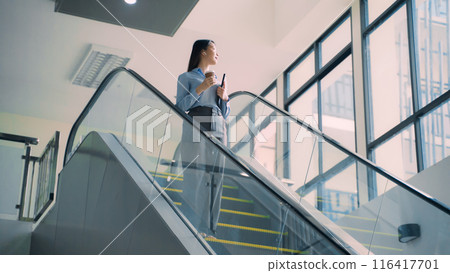 A business woman is walking down to escalator with a coffee cup in her hand A business woman is walking down to escalator with a coffee cup in her hand 116417701