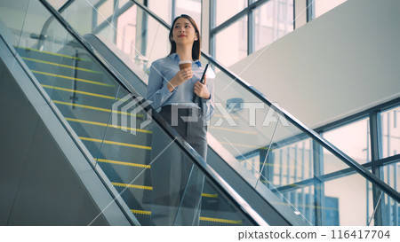 A business woman is walking down to escalator with a coffee cup in her hand 116417704