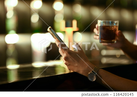 Close up of female hand holding a glass of whisky and using mobile phone at the bar counter 116420145