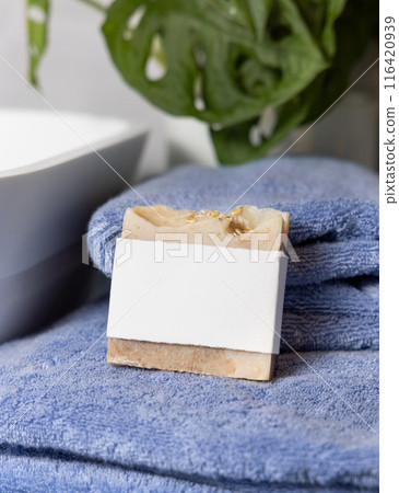 Soap bar with blank label on blue towels near vessel sink and green plant close up, mockup 116420939