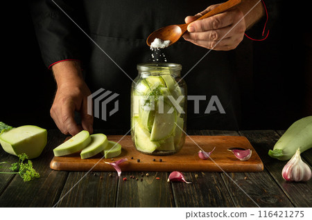 A cook spoons salt into a jar of chopped vegetable marrow before canning it with dill and garlic. Concept of pickling delicious zucchini on the kitchen table A cook spoons salt into a jar of chopped vegetable marrow before canning it with dill and garlic. Concept of pickling delicious zucchini on the kitchen table 116421275