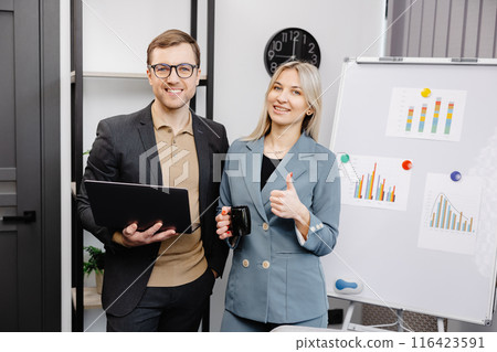 Business people with laptop discussing new projects in office. A young woman and a man stand near flipcharts and solve work issues Business people with laptop discussing new projects in office. A young woman and a man stand near flipcharts and solve work issues 116423591