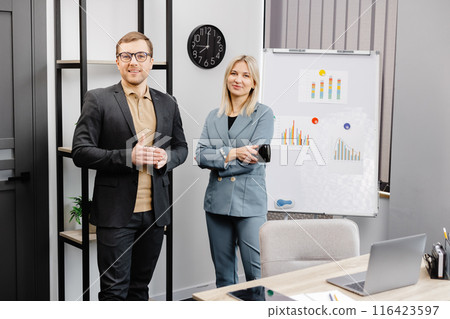 Couple of cheerful colleagues talking while standing at the flipchart in office with cups of coffee Couple of cheerful colleagues talking while standing at the flipchart in office with cups of coffee 116423597
