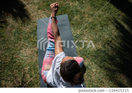 Woman practicing yoga outdoors on a mat, wearing colorful leggings and white top, performing a seated twist posture in a sunny garden 116425215