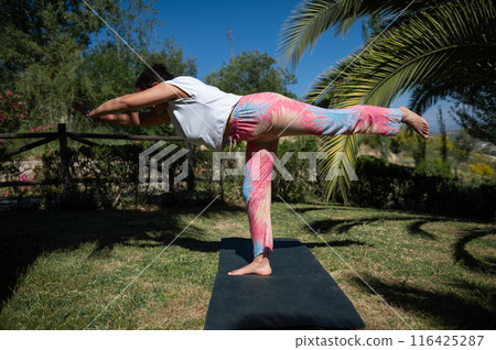 Woman practicing yoga outdoors, balancing on one leg in warrior pose on a sunny day, wearing colorful pants Woman practicing yoga outdoors, balancing on one leg in warrior pose on a sunny day, wearing colorful pants 116425287