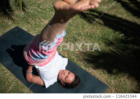 Woman practicing yoga outdoors on a sunny day, performing a leg lift exercise on a mat 116425344