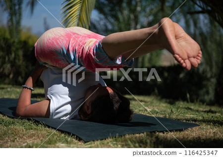 Woman practicing yoga outdoors on a sunny day, performing shoulder stand pose on mat in nature Woman practicing yoga outdoors on a sunny day, performing shoulder stand pose on mat in nature 116425347