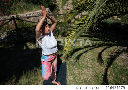 Woman practicing yoga outdoors in nature, performing tree pose with closed eyes under palm trees 116425379