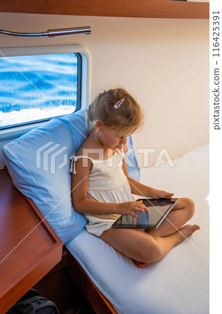 Little girl sits with tablet near window in the cabin of a yacht during a sea crossing. Yacht life concept Little girl sits with tablet near window in the cabin of a yacht during a sea crossing. Yacht life concept 116425391