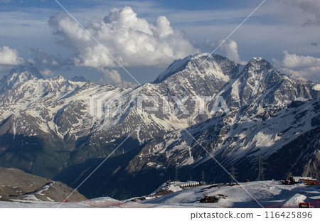 Caucasus Range at sunset with snow-capped peaks, clouds hover over rugged terrain. Alpine landscape in soft evening light, tranquil mountaintop scene attracts nature lovers, hikers. 116425896