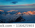Golden sunset lights snowy peaks of Caucasus Range. Majestic mountain landscape glow at dusk. Clear blue sky, white clouds. Nature beauty, travel destination for hikers, photographers. 116425903