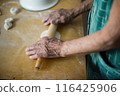 Elderly woman rolls dough for vareniki on kitchen table. Traditional Ukrainian cuisine, homemade pierogi making process. Granny prepares authentic Eastern European dish, culinary skills displayed. 116425906