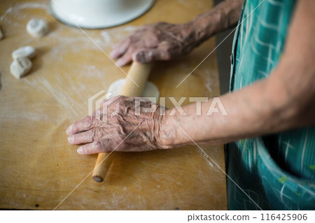 Elderly woman rolls dough for vareniki on kitchen table. Traditional Ukrainian cuisine, homemade pierogi making process. Granny prepares authentic Eastern European dish, culinary skills displayed. Elderly woman rolls dough for vareniki on kitchen table. Traditional Ukrainian cuisine, homemade pierogi making process. Granny prepares authentic Eastern European dish, culinary skills displayed. 116425906