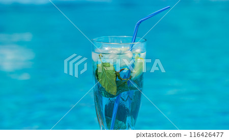 A refreshing drink with mint and ice in a glass with a blue straw against a bright blue water background. A refreshing drink with mint and ice in a glass with a blue straw against a bright blue water background. 116426477