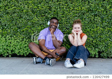African man and Caucasian woman are sitting on ground with background full of green plant, both wearing headphones. They are smiling and seem to be enjoying their music African man and Caucasian woman are sitting on ground with background full of green plant, both wearing headphones. They are smiling and seem to be enjoying their music 116427499