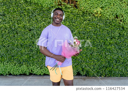 Happy African man with headphones wearing a purple shirt is holding a bouquet of flowers. He is wearing headphones and he is listening to music Happy African man with headphones wearing a purple shirt is holding a bouquet of flowers. He is wearing headphones and he is listening to music 116427534