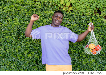 Happy healthy sporty African man in a purple shirt holding reusable cotton mesh bag with fruit on green plant wall background. Happy healthy sporty African man in a purple shirt holding reusable cotton mesh bag with fruit on green plant wall background. 116427553