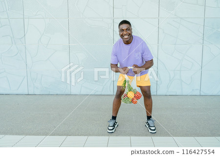 Strong and sporty African hipster man stands against wall carrying vegetables in reusable cotton mesh shopping bag, living green lifestyle. Strong and sporty African hipster man stands against wall carrying vegetables in reusable cotton mesh shopping bag, living green lifestyle. 116427554