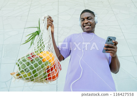Ordering groceries online concept. Wide angle, bottom view of African man standing spending days using reusable cotton mesh bag and smart phone. Ordering groceries online concept. Wide angle, bottom view of African man standing spending days using reusable cotton mesh bag and smart phone. 116427558