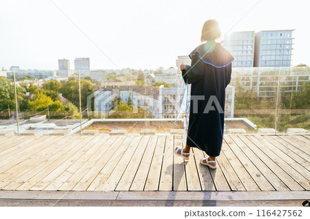 Back view unrecognizable woman wearing ceremony robe holding certificate with city view on background. Girl celebrating graduation and getting diploma. Education, graduation and people concept. Back view unrecognizable woman wearing ceremony robe holding certificate with city view on background. Girl celebrating graduation and getting diploma. Education, graduation and people concept. 116427562