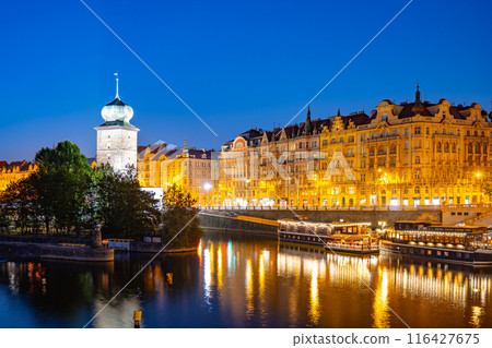 The Sitkov Water Tower stands illuminated on the Masaryk Embankment in Prague, Czechia, as dusk descends. The tower is reflected in the tranquil waters of the Vltava River. 116427675