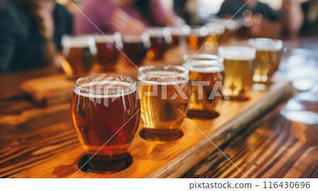 A row of assorted beers in a tasting flight, displayed on a wooden table at a bustling brewery. 116430696