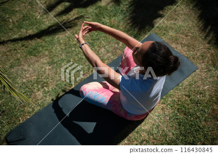 Woman practicing yoga outdoors on a sunny day, seated on a yoga mat in a peaceful garden setting 116430845