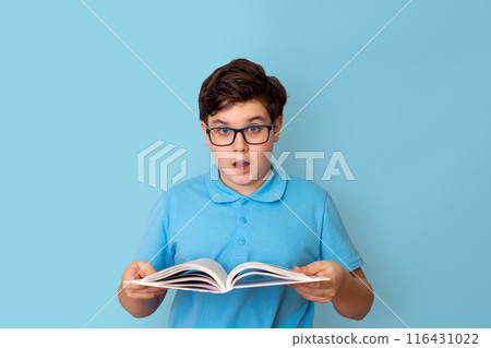 A surprised schoolboy in glasses, holding an open book in his hands, look at camera 116431022