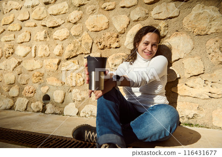 Woman sitting outdoors with coffee cup against stone wall 116431677