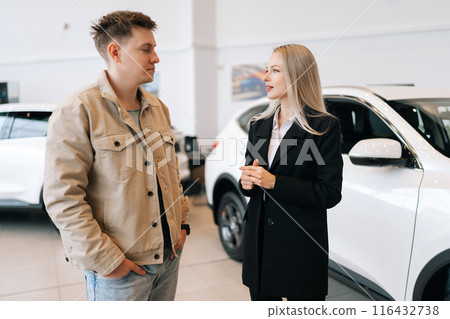 Portrait of attractive blonde female manager in suit showing new cars to male buyer and walking in modern showroom spbas. Young woman car dealer talking to man client and showing models of transport. Portrait of attractive blonde female manager in suit showing new cars to male buyer and walking in modern showroom spbas. Young woman car dealer talking to man client and showing models of transport. 116432738