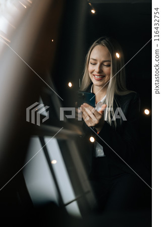 Vertical portrait through windshield to cheerful female passenger reading good news using smartphone sitting on front seat in car. Pretty young woman browsing phone, surfing internet, texting sms. 116432754