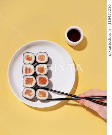 A hand holds chopsticks with Japanese sushi rolls on a white plate and soy sauce  116433256