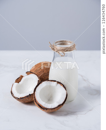 Glass vegan milk bottle on white marble table with half coconuts. Front view and copy space 116434760