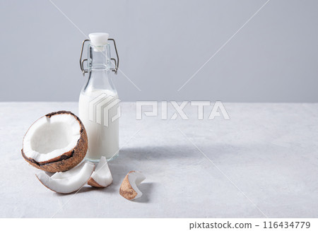Glass vegan milk bottle on white marble table with half coconuts. Front view and copy space 116434779