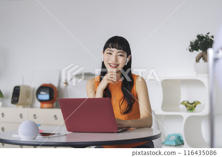 Young woman wearing earphones and sitting in front of a laptop looking at the camera 116435086