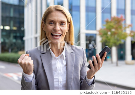 Confident businesswoman in gray suit celebrating success while using smartphone outside modern office building. Concept of business achievement, joy, and professional success. Confident businesswoman in gray suit celebrating success while using smartphone outside modern office building. Concept of business achievement, joy, and professional success. 116435155