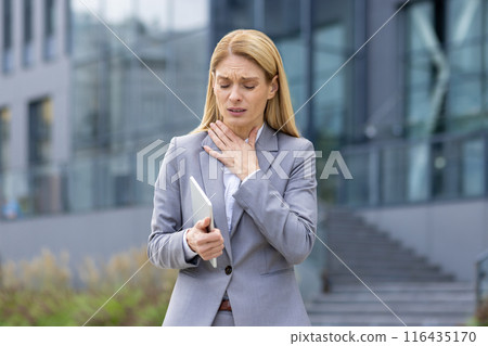 Businesswoman in gray suit holding tablet outside modern office building, experiencing throat discomfort. Scene portrays stress, pain, and health issues in a business environment. 116435170
