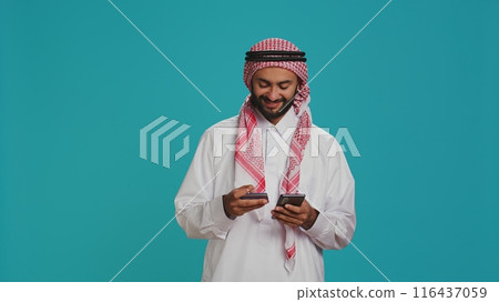 Joyful model using card to make payment while shopping on clothing store website using smartphone app. Middle eastern adult in islamic costume doing banking transfer to buy on sale. Joyful model using card to make payment while shopping on clothing store website using smartphone app. Middle eastern adult in islamic costume doing banking transfer to buy on sale. 116437059